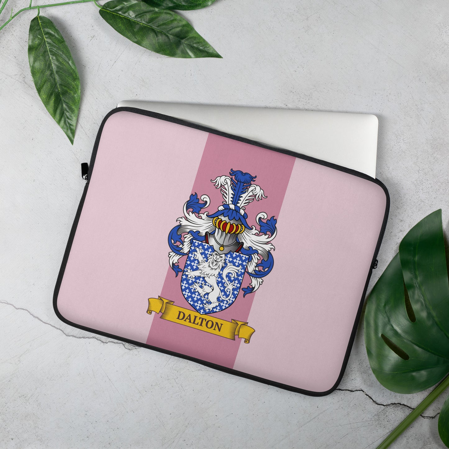 Laptop sleeve with a coat of arms design on a pink background, placed on a light surface with green leaves.